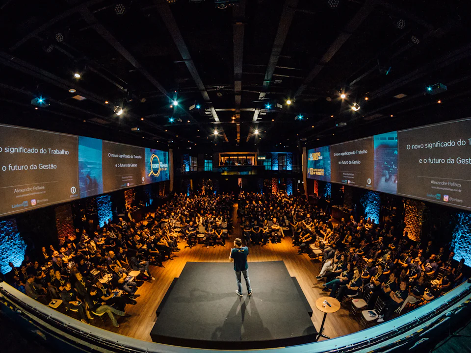 A speaker on a black stage in the middle of a full auditorium, screens on both side walls