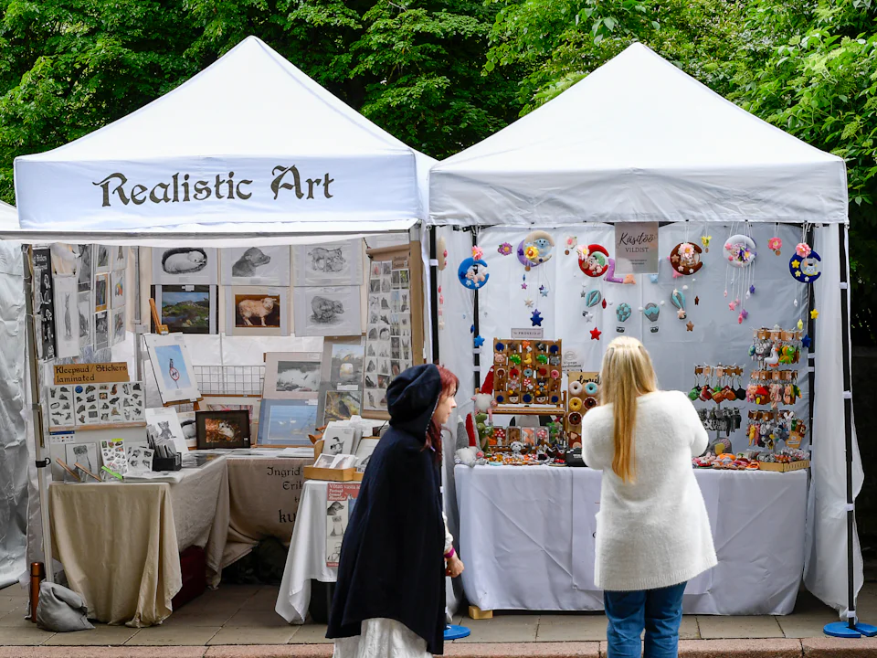 Two visitors browsing drawings and handmade crafts under two white tents at an outdoor market