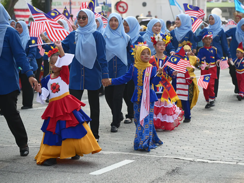 Children in traditional costume and teachers in blue marching with Malaysian flags in a National Day parade