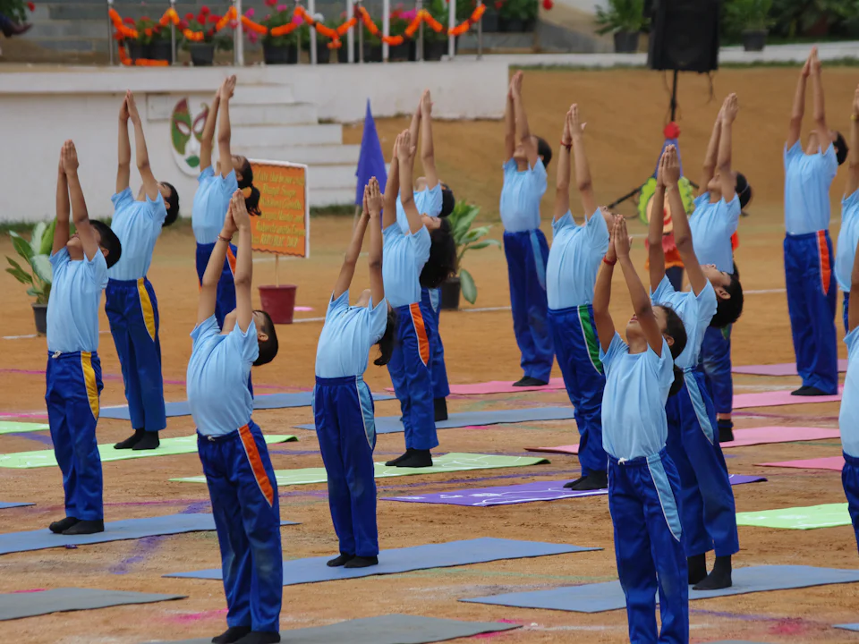 Rows of students in blue school tracksuits stretching their arms overhead on a sports field